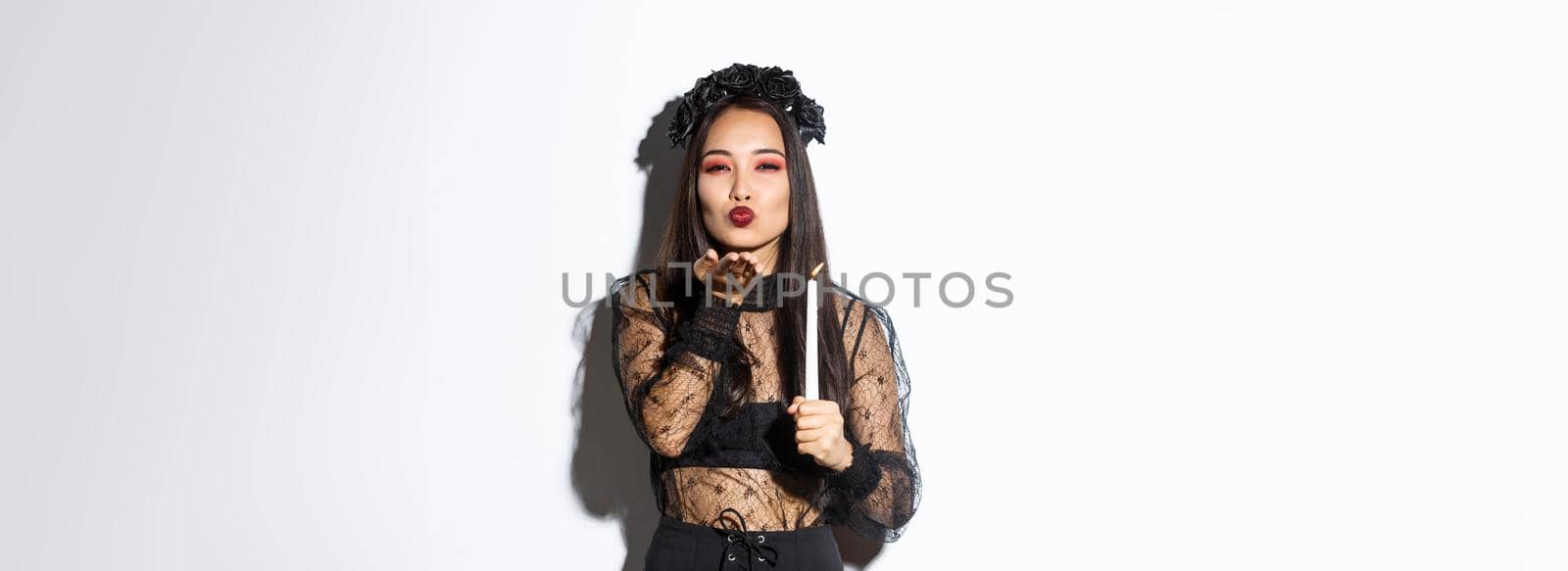 Beautiful and elegant asian woman in gothic lace dress celebrating halloween, dress-up as witch, sending air kiss at camera and holding candle.