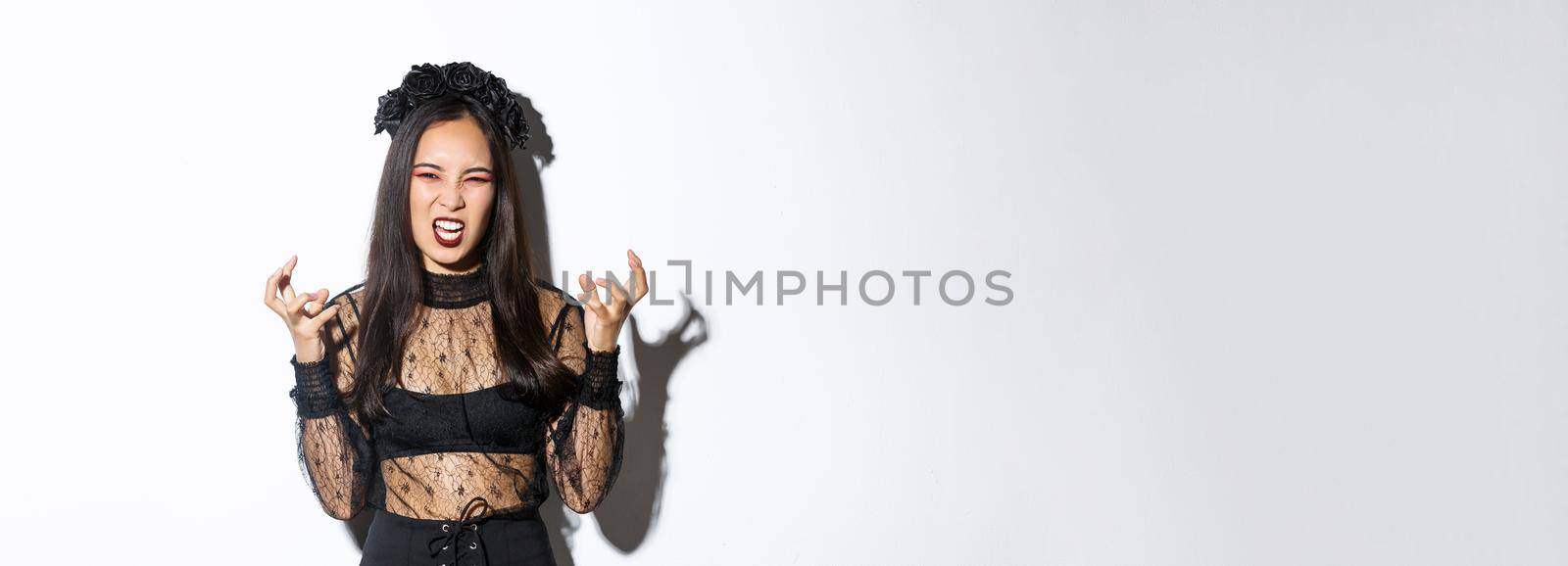 Image of woman in halloween costume looking like angry evil witch, cursing and clenching fists mad. Female in black dress and wreath trick or treating, standing white background by Benzoix