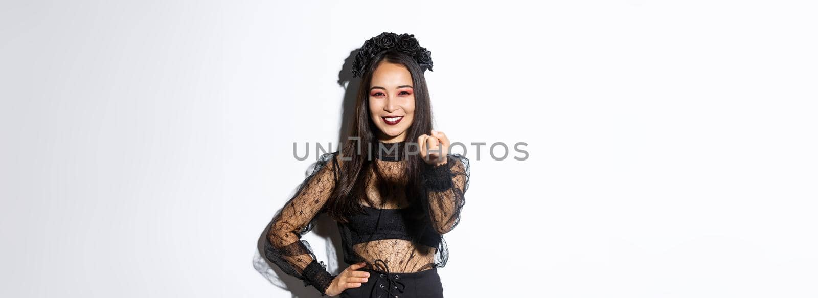 Beautiful woman in witch costume telling to come closer, flick finger and smiling, standing in halloween outfit, gothic makeup, standing over white background by Benzoix