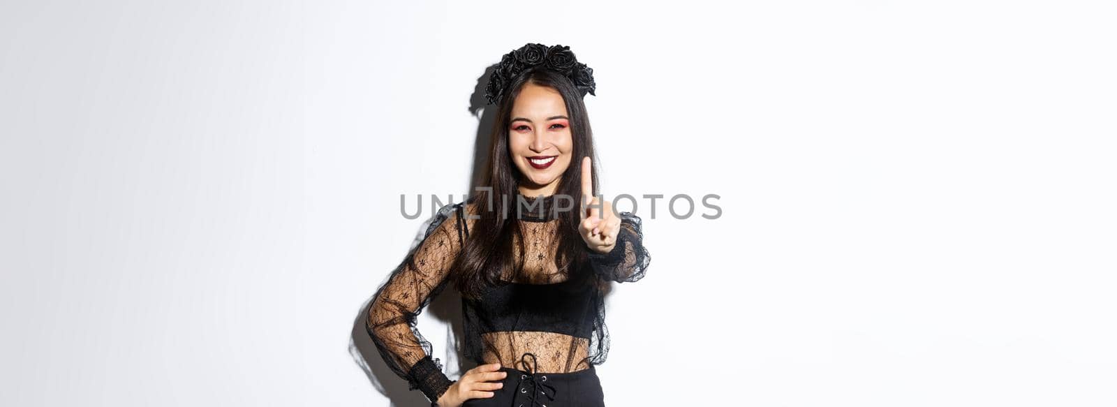 Image of smiling beautiful asian woman in halloween costume showing stop gesture, extend one finger and looking happy, prohibit or reject something, standing white background by Benzoix