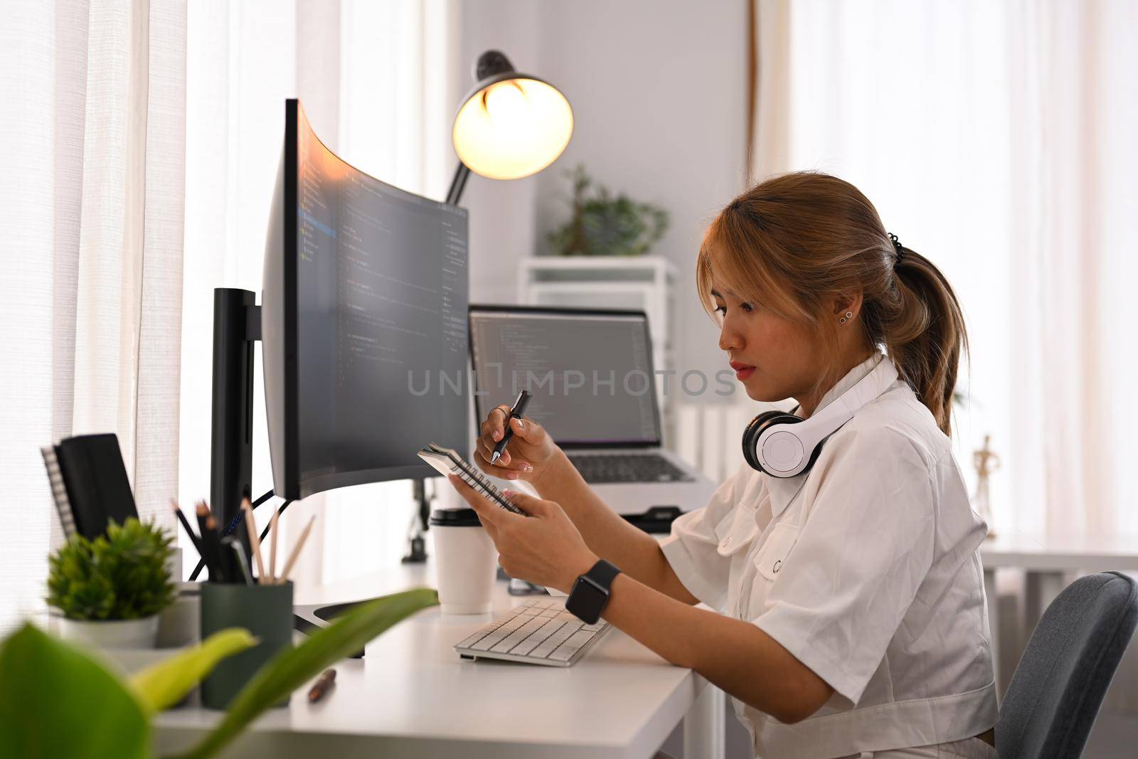 Young female programmer sitting front of computers with coded data on screen and making notes on notebook.