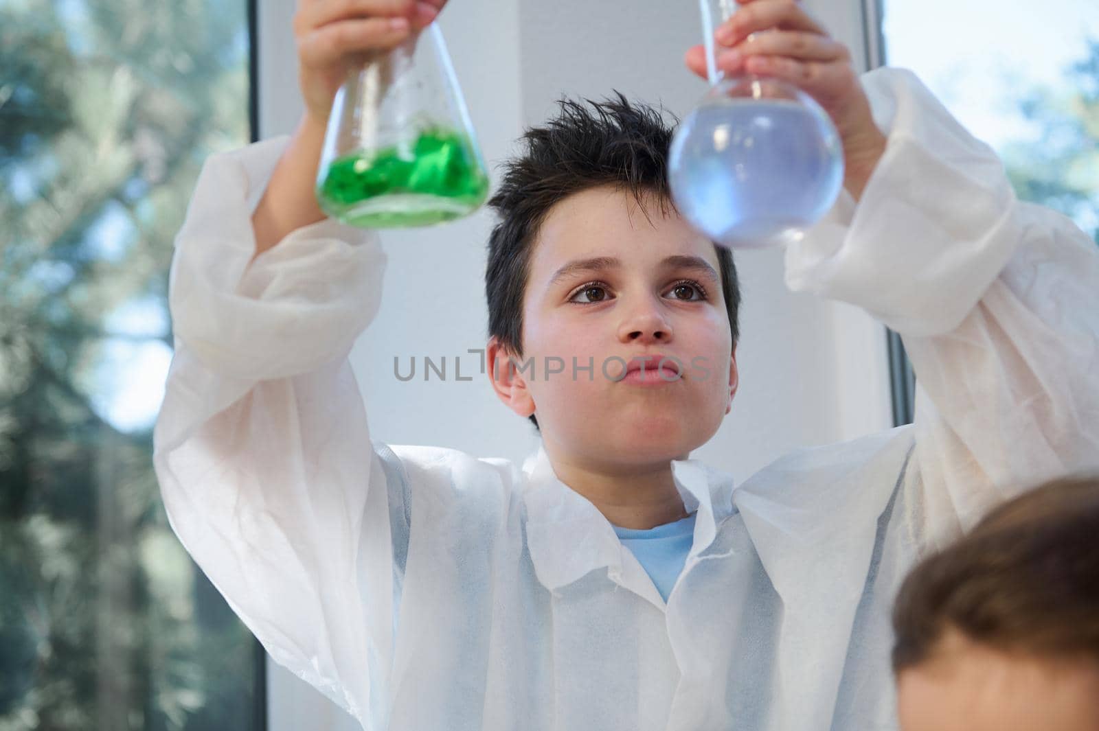 Handsome Caucasian school boy, a scientist chemist in a white lab coat, holding glass laboratory flasks with chemicals, watching the reaction taking place between liquid and reagents. Chemistry class.