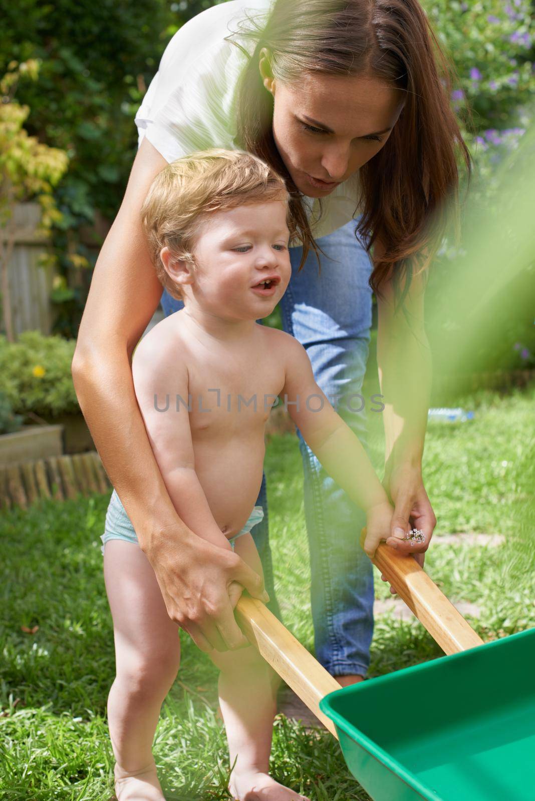 Learning about gardening. An infant boy pushing his toy wheel barrow while his mother helps