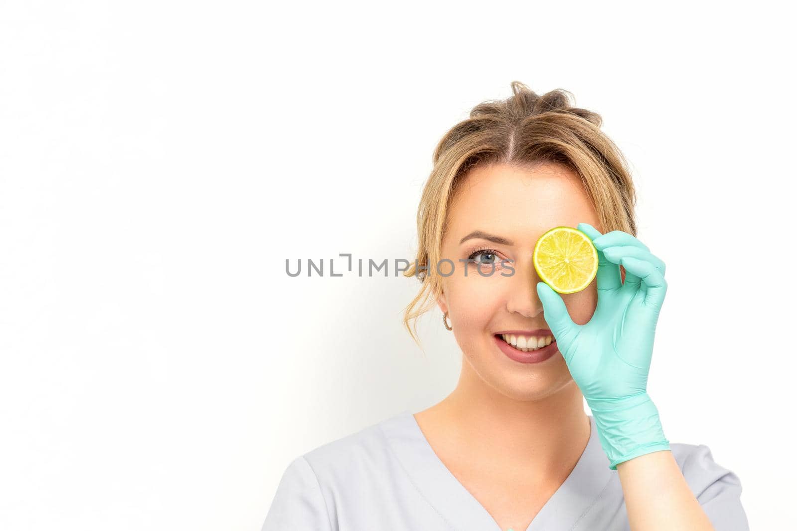 Portrait of young caucasian smiling female beautician covering eye with a lime slice wearing gloves over a white background