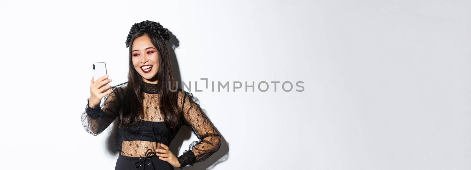 Portrait of happy beautiful asian woman in halloween costume smiling and looking at mobile phone screen, having video call, standing over white background by Benzoix