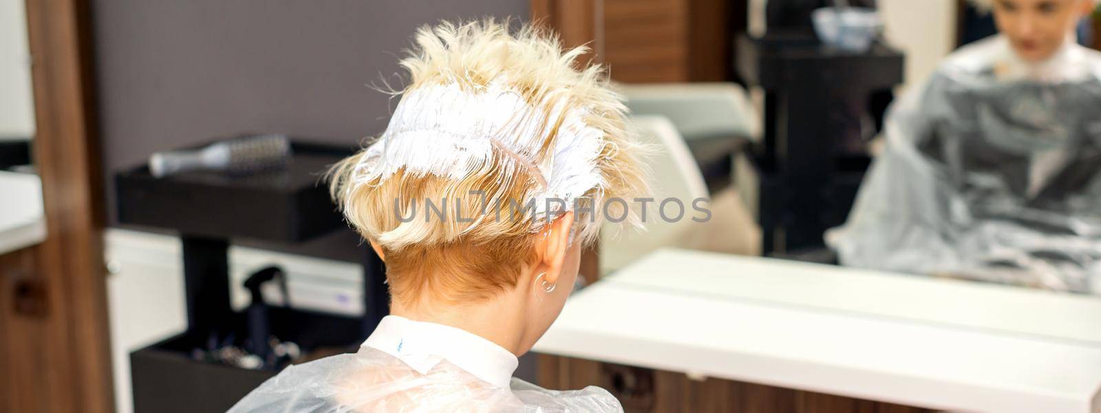 Coloring white hair with hair dye of the young caucasian blonde woman sitting at a hair salon, close up
