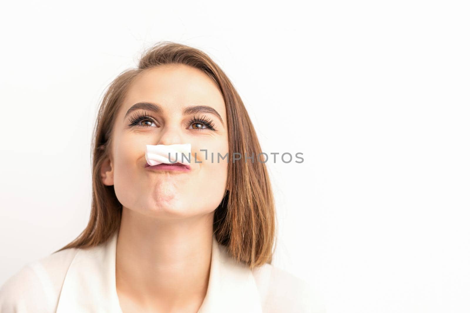 Funny young woman with marshmallow on her lips looking up and standing on white background with copy space