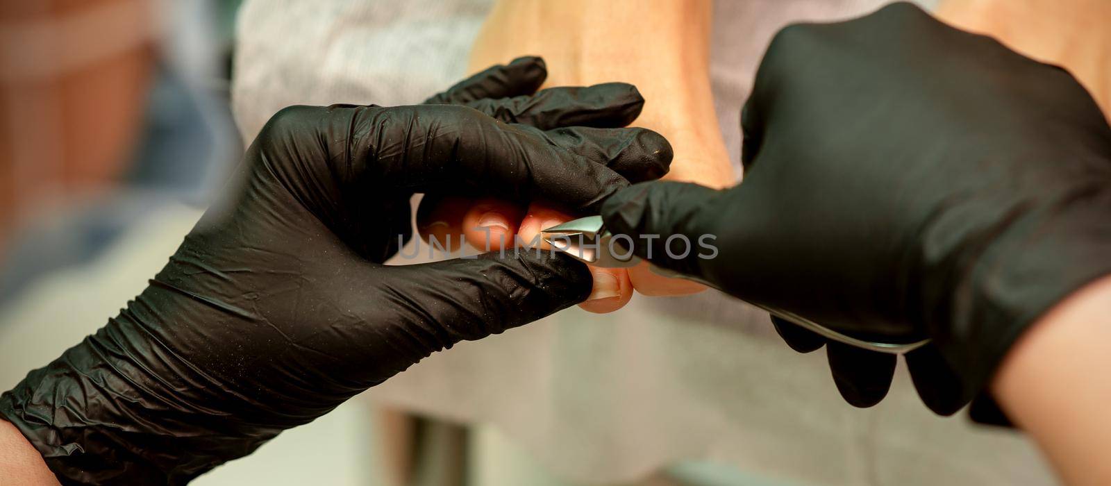 Professional pedicure. Pedicure master wearing latex gloves cuts female toenails in the beauty salon, closeup. by okskukuruza