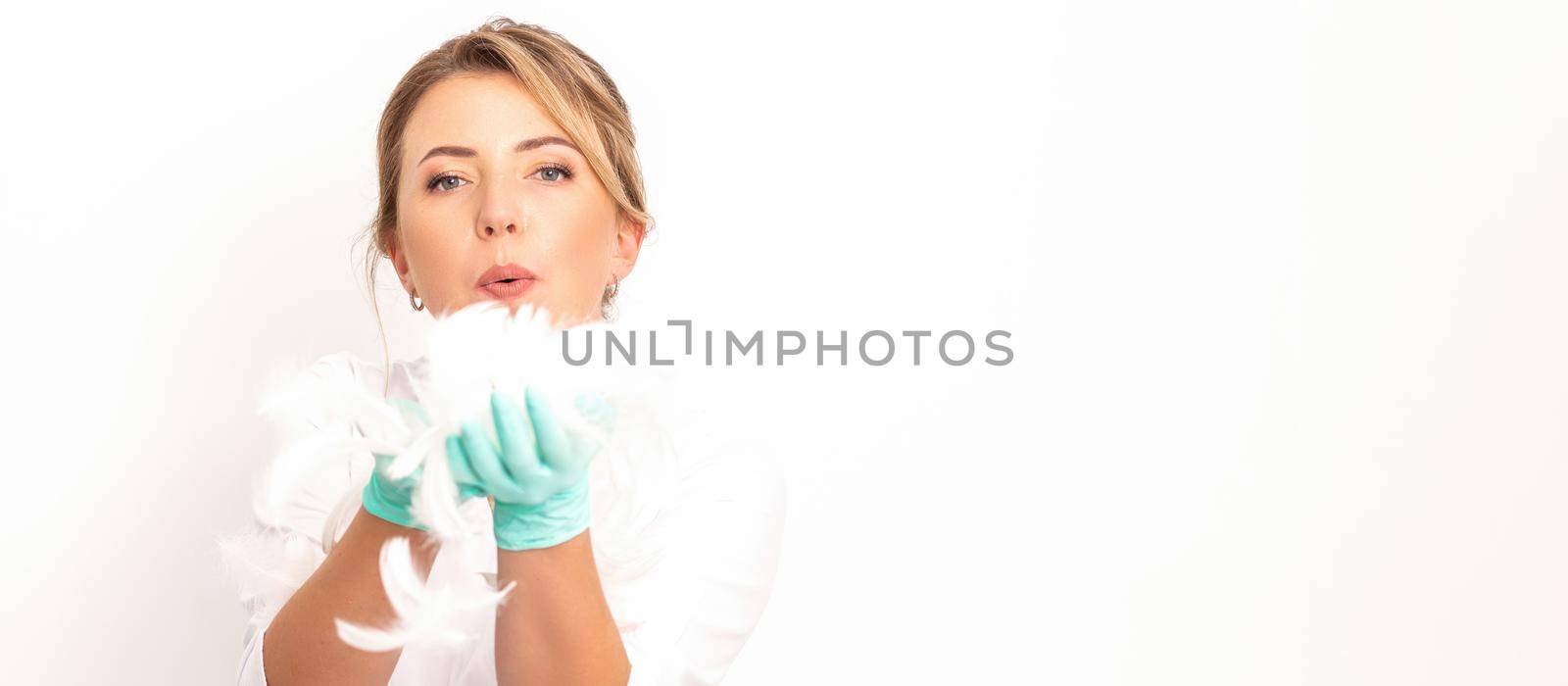 Young beautiful woman beautician in protective green gloves standing and blowing on feathers over white wall background. by okskukuruza