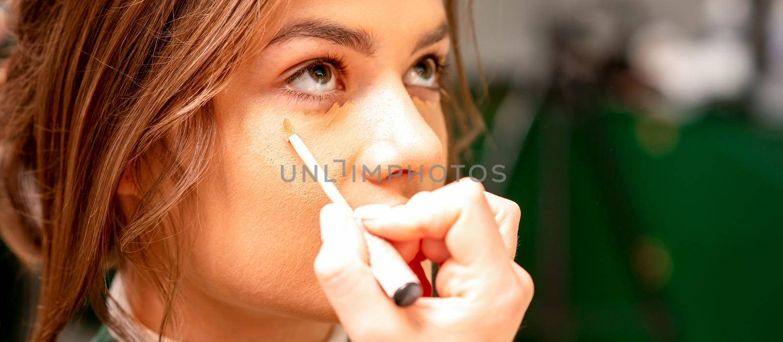 Beautiful young brunette woman receiving makeup with stick concealer on her face in a beauty salon