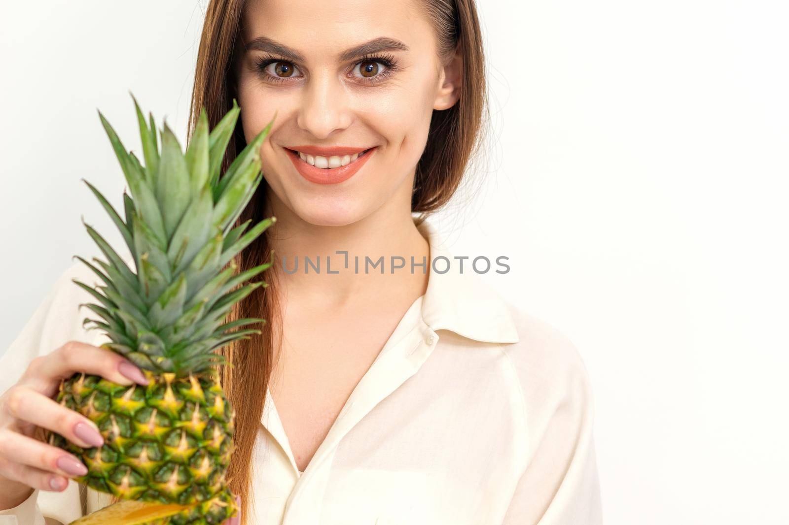 Beautiful young Caucasian woman holding pineapple and smiling, wearing a white shirt over white background. by okskukuruza