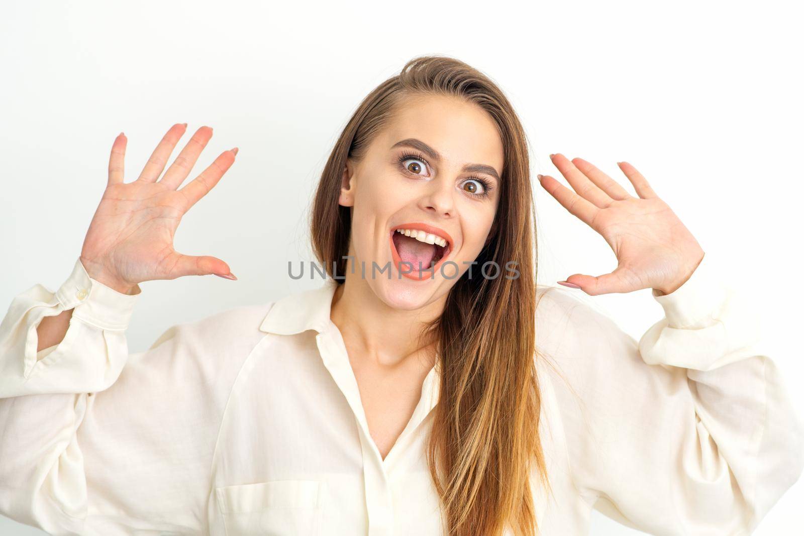 Portrait of young caucasian woman wearing white shirt raises hands and laughs positively with open mouth poses against a white background