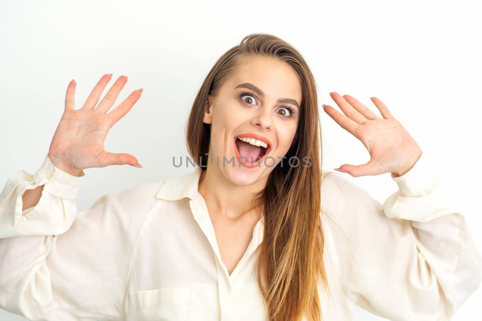 Portrait of young caucasian woman wearing white shirt raises hands and laughs positively with open mouth poses against a white background. by okskukuruza