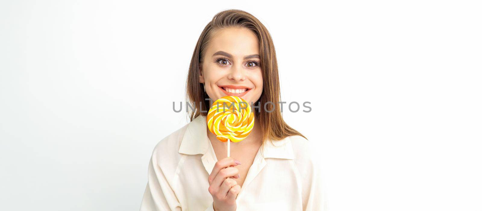 Beautiful young caucasian woman wearing a white shirt licking a lollipop on a white background