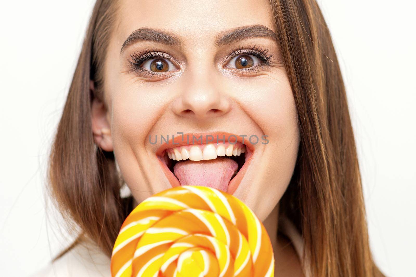 Beautiful young caucasian woman wearing a white shirt licking a lollipop on a white background