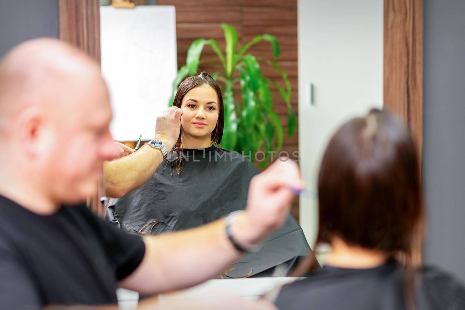 Reflection in the mirror of the young caucasian woman sitting and receiving haircut by male hairdresser at hairdresser salon. by okskukuruza