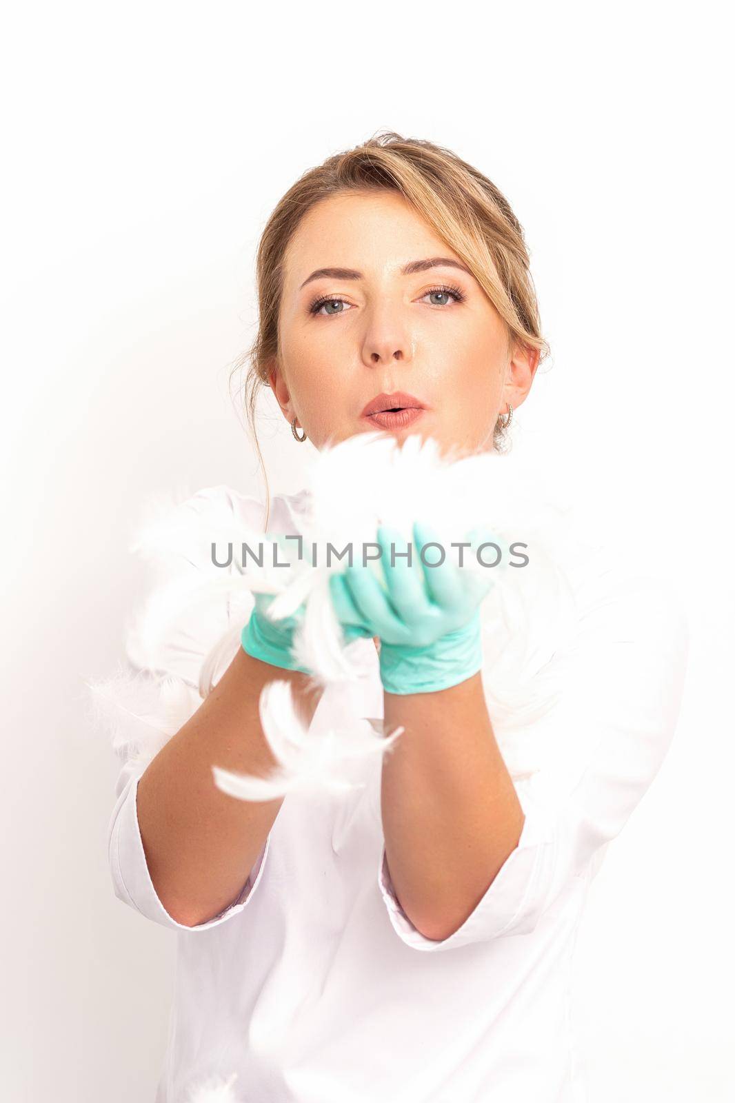 Young beautiful woman beautician in protective green gloves standing and blowing on feathers over white wall background