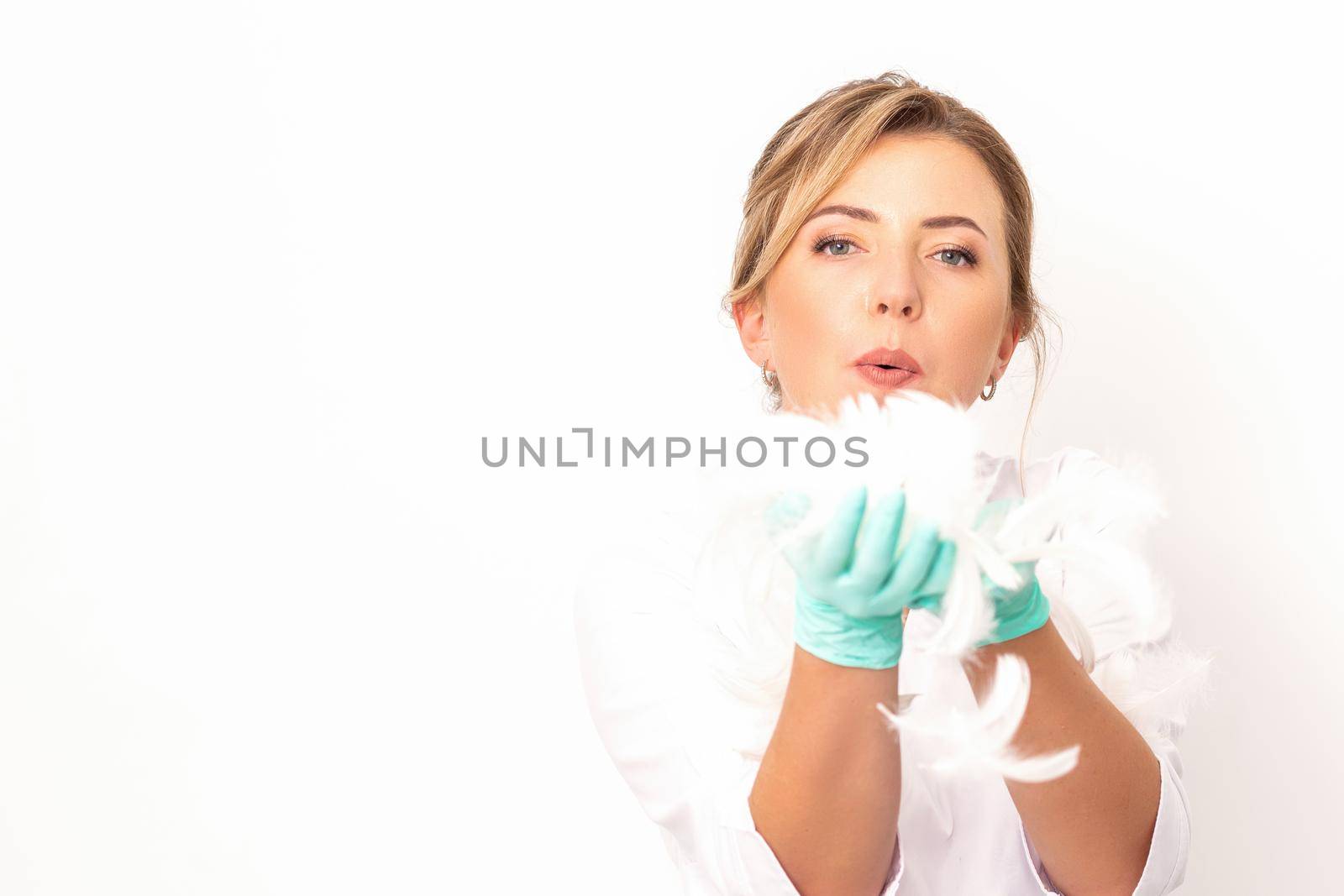 Young beautiful woman beautician in protective green gloves standing and blowing on feathers over white wall background