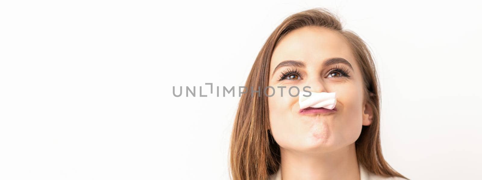 Funny young woman with marshmallow on her lips looking up and standing on white background with copy space