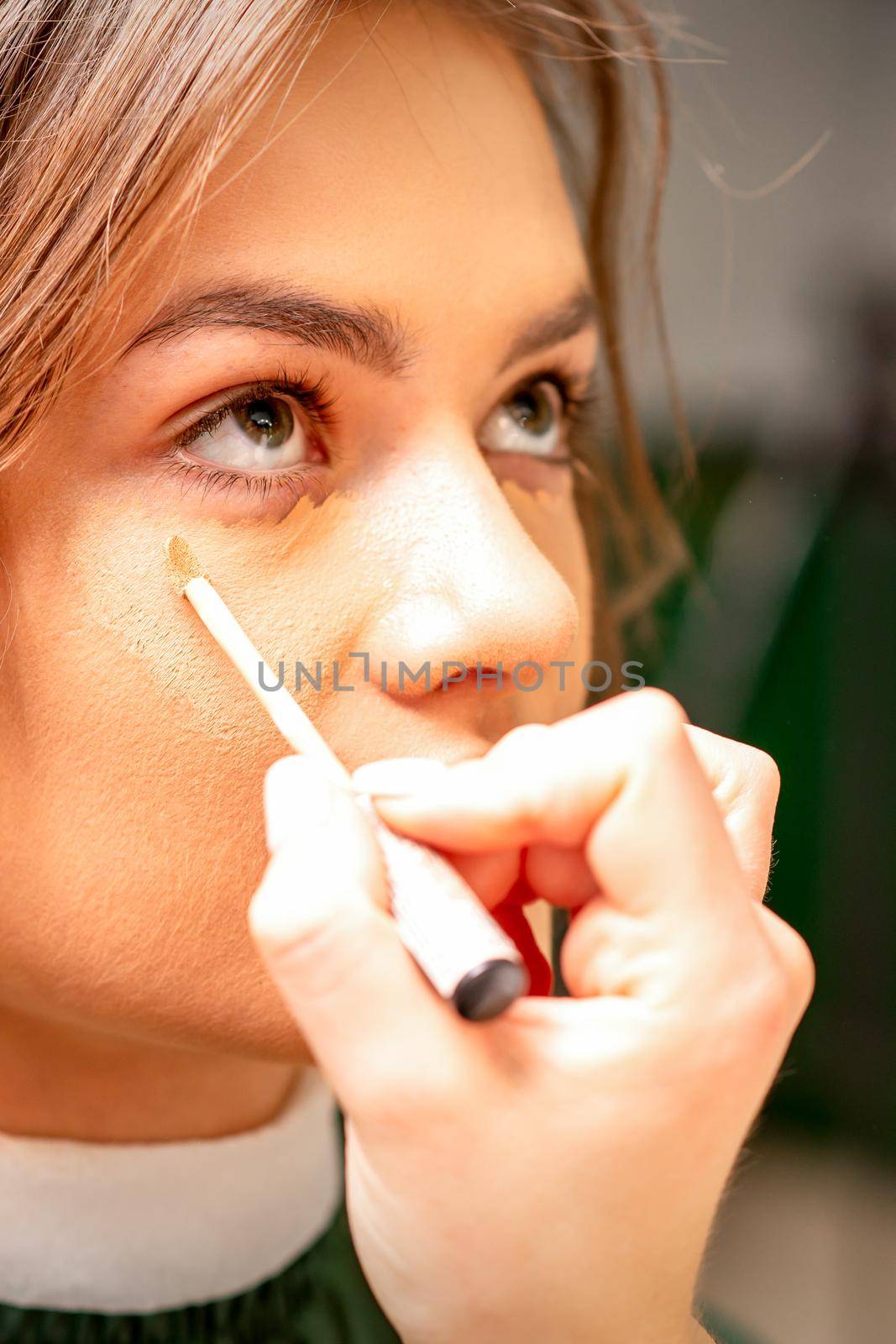 Beautiful young brunette woman receiving makeup with stick concealer on her face in a beauty salon