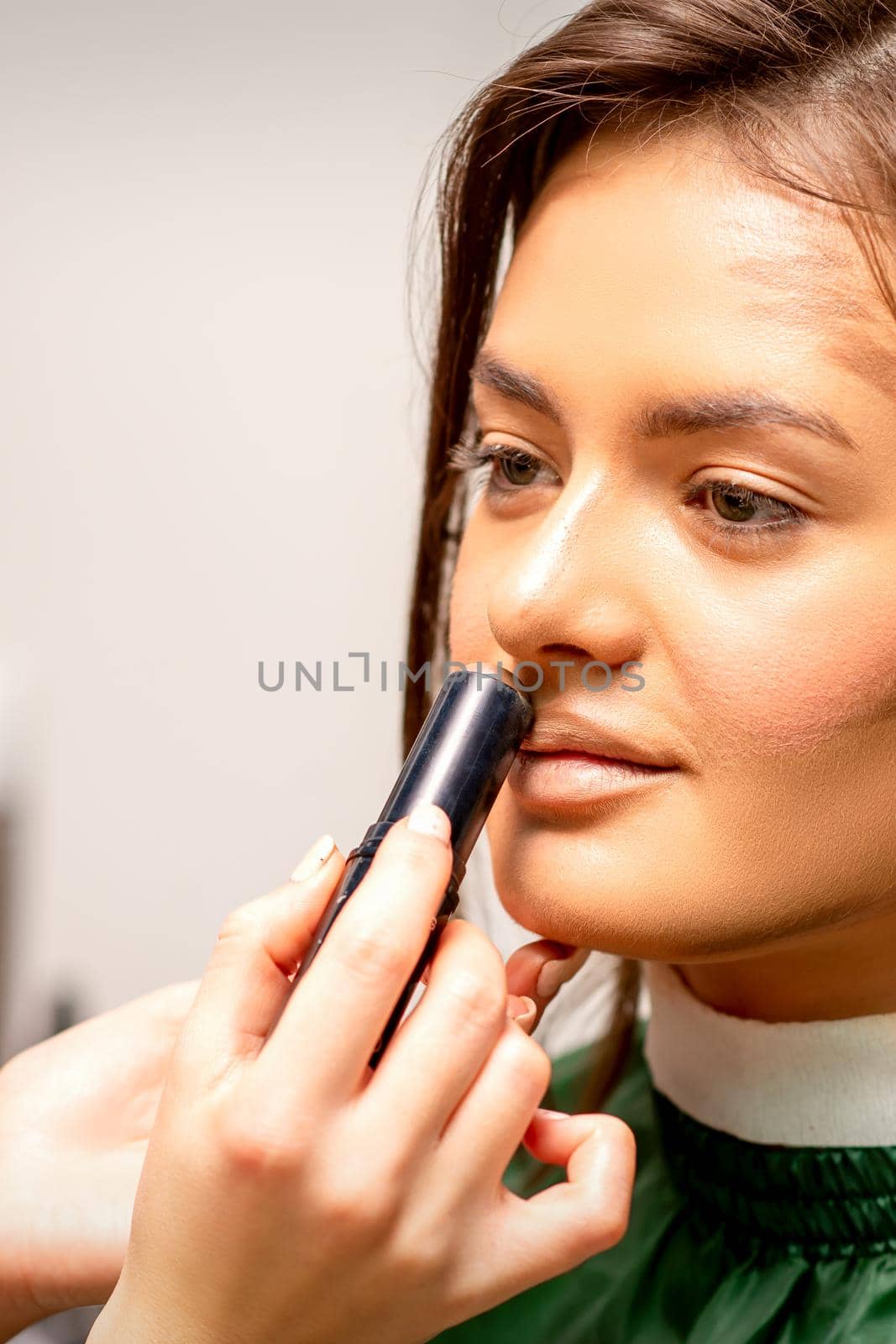 Beautiful young brunette woman receiving makeup with stick concealer on her face in a beauty salon