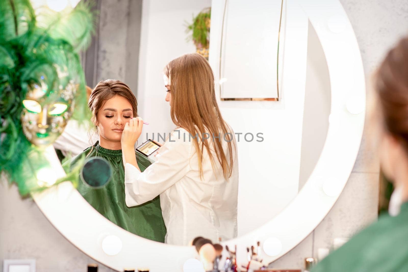 Professional makeup artist applies makeup to the eyes of the beautiful young woman in the mirror smiling in a beauty salon