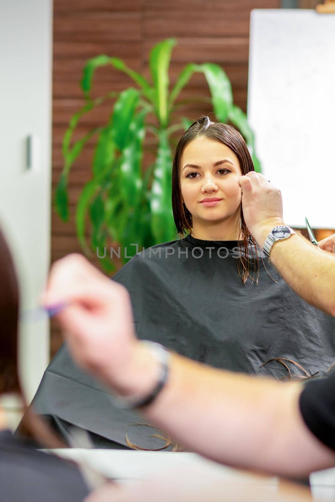 Reflection in the mirror of the young caucasian woman sitting and receiving haircut by male hairdresser at hairdresser salon