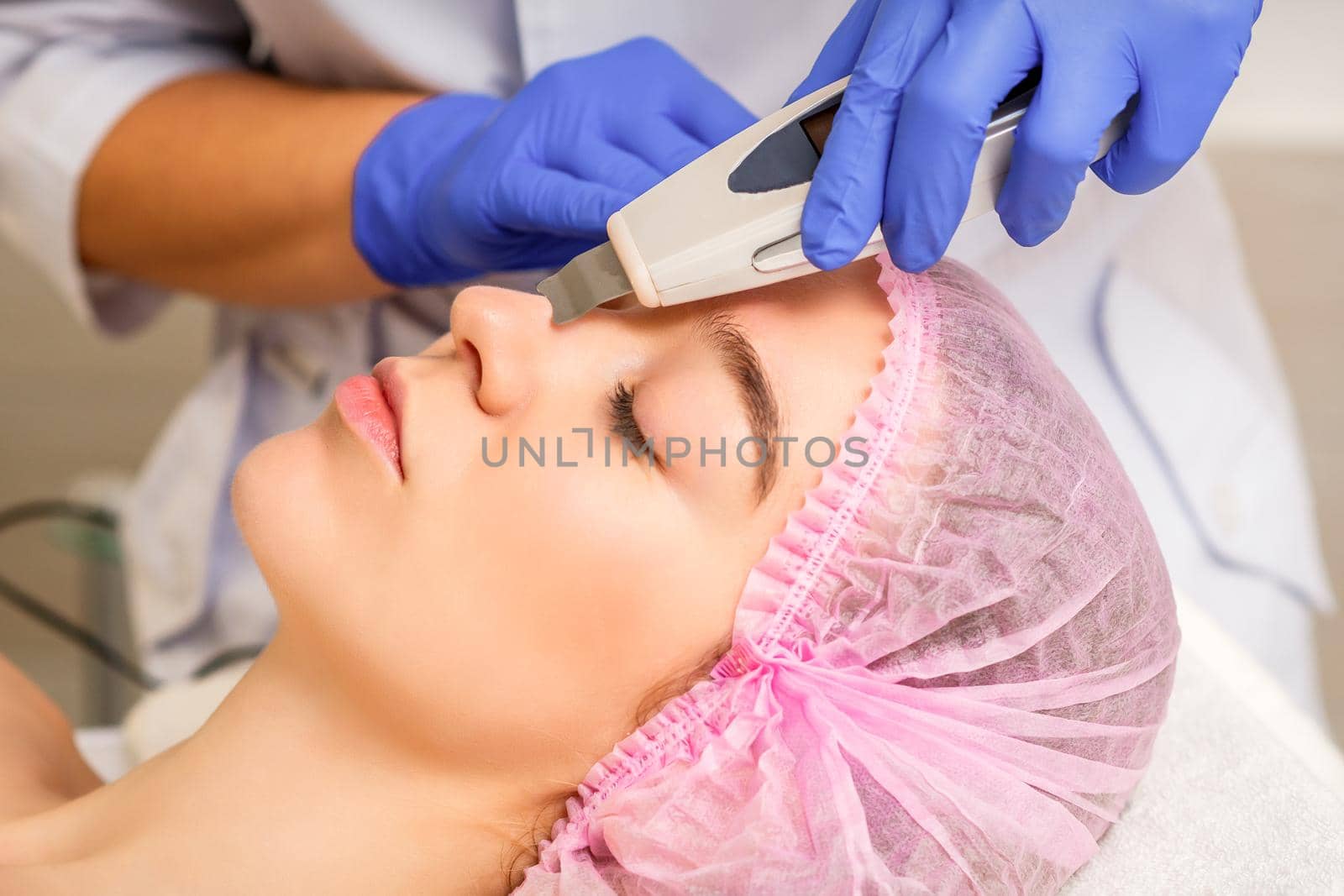 Young caucasian woman receiving facial skin cleaning by ultrasonic cosmetology face equipment in a medical salon