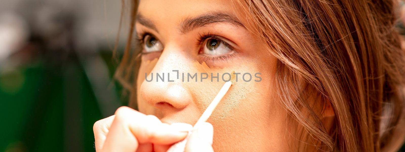 Beautiful young brunette woman receiving makeup with stick concealer on her face in a beauty salon. by okskukuruza
