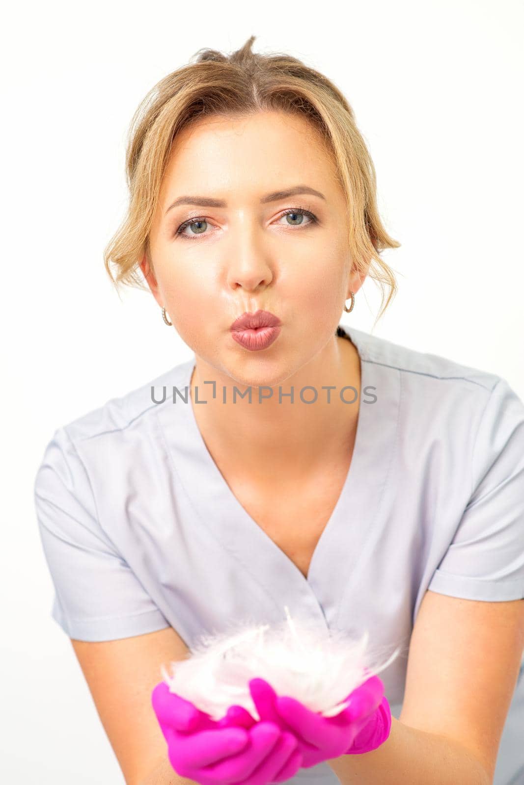 Young beautiful woman beautician in protective pink gloves standing and holding white feathers over white wall background