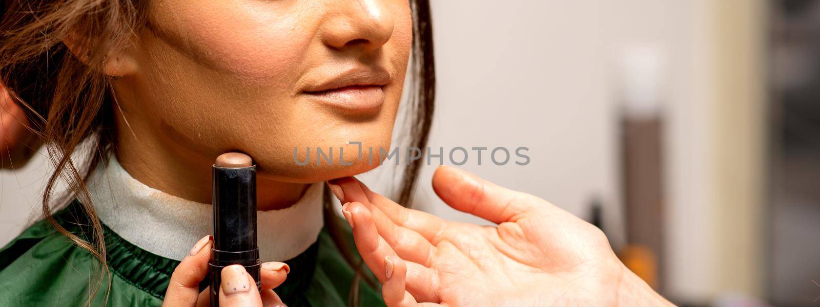 Beautiful young brunette woman receiving makeup with stick concealer on her face in a beauty salon. by okskukuruza