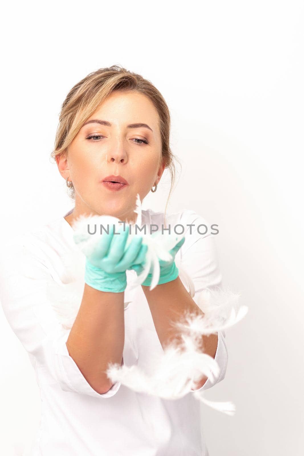 Young beautiful woman beautician in protective green gloves standing and blowing on feathers over white wall background