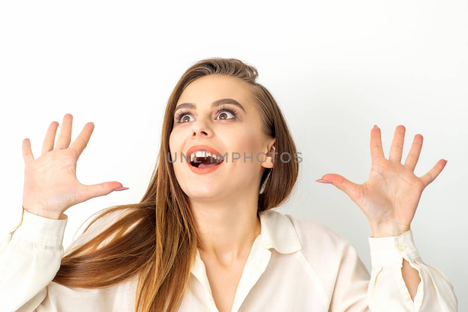 Portrait of young caucasian woman wearing white shirt raises hands and laughs positively with open mouth looking up against a white background