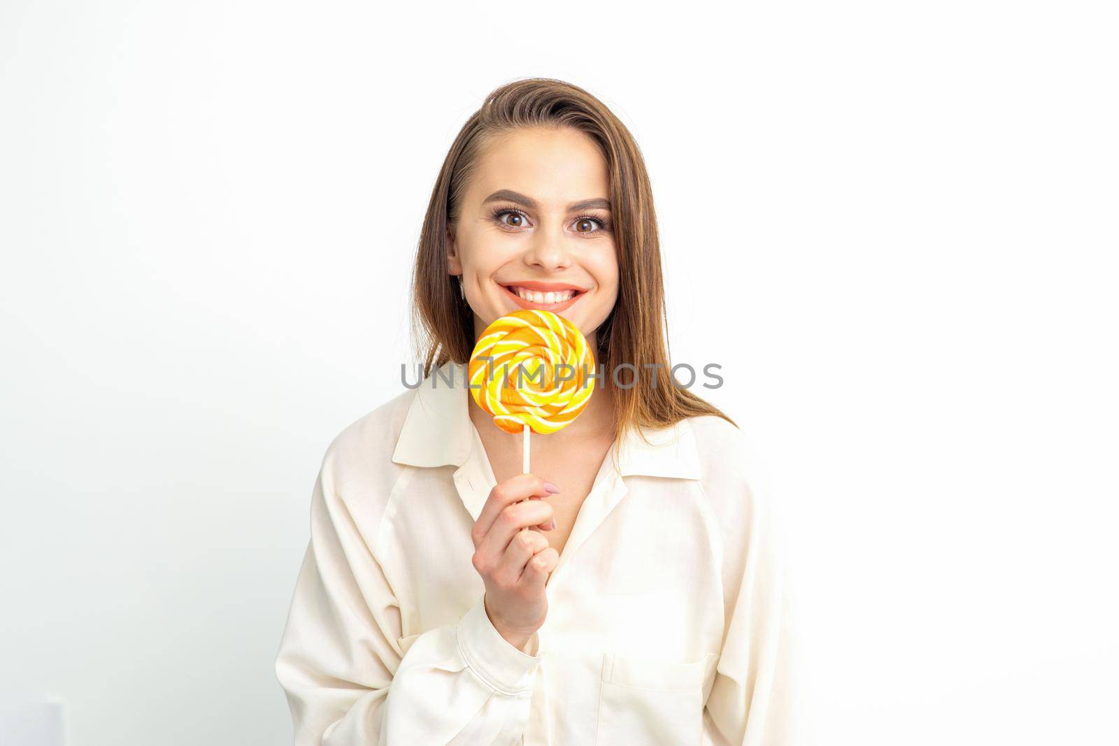 Beautiful young caucasian woman wearing a white shirt licking a lollipop on a white background. by okskukuruza