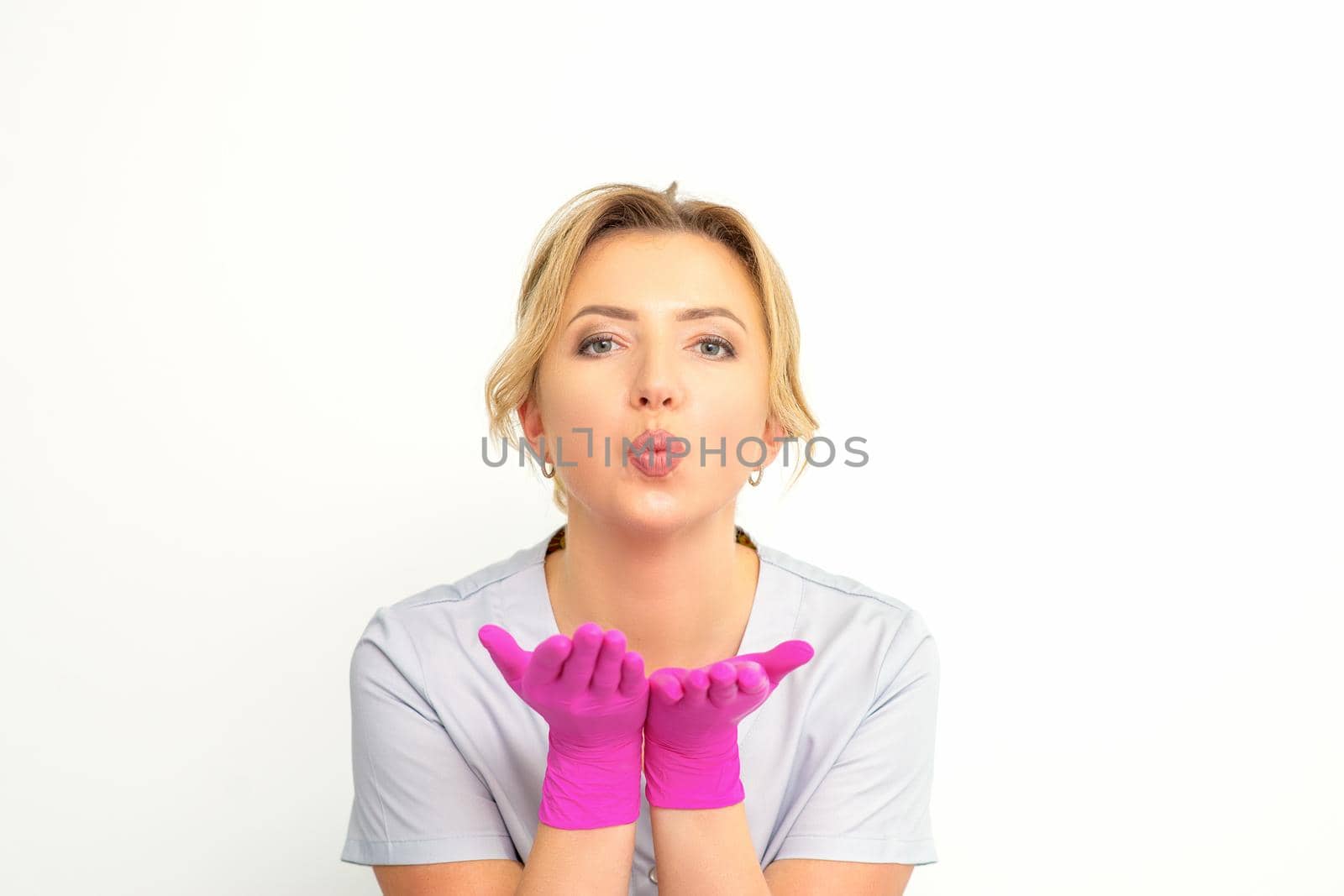 Young Caucasian doctor woman wearing pink gloves folding lips and holding palms to send air kiss isolated white background. by okskukuruza