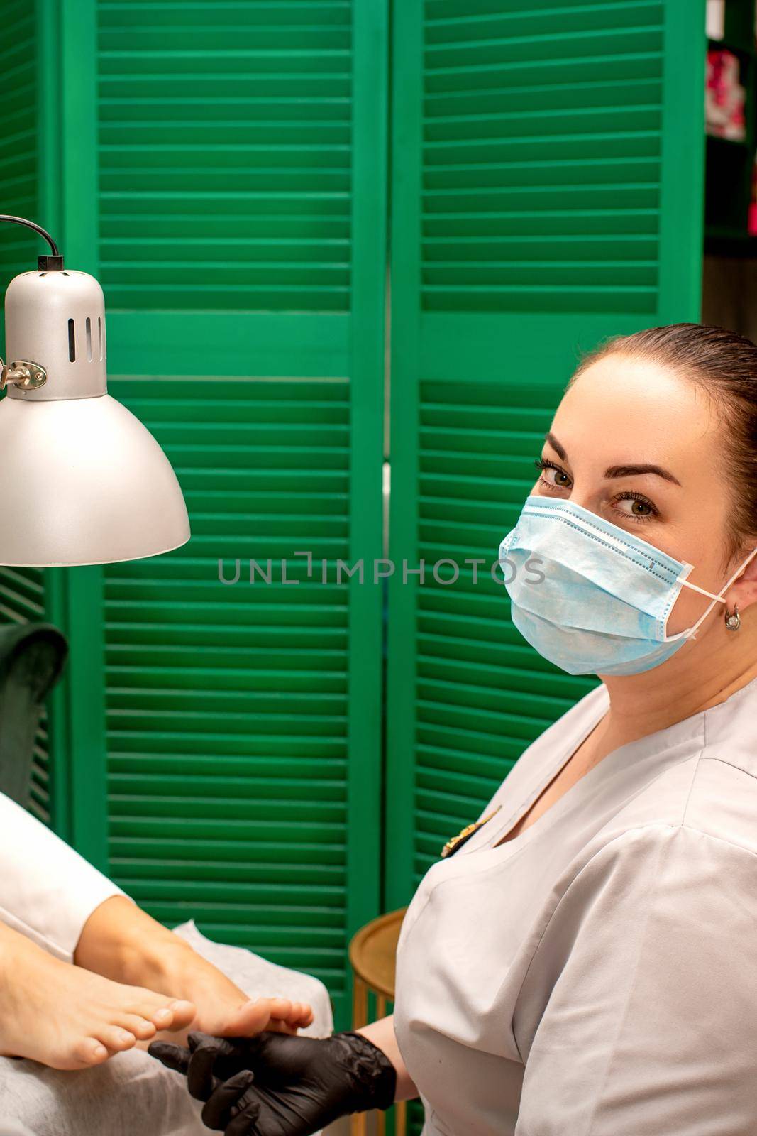 Young female podiatrist with a protective mask doing chiropody and looking at the camera in her podiatry clinic. by okskukuruza