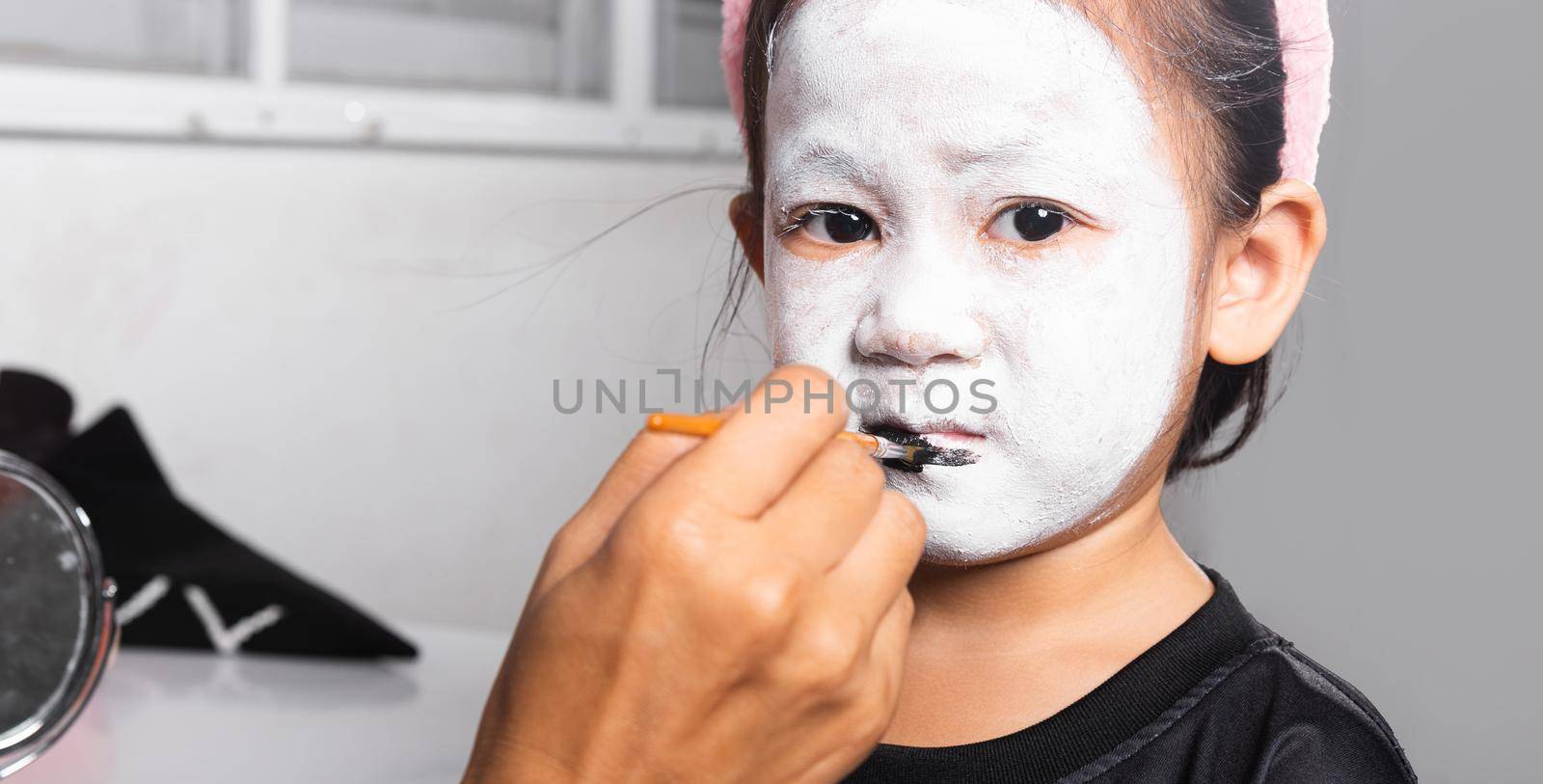 Asian mother and kid daughter getting makeup halloween face painting look like ghost, Portrait of woman applying color brush to child girl face for party at home, Happy halloween day concept
