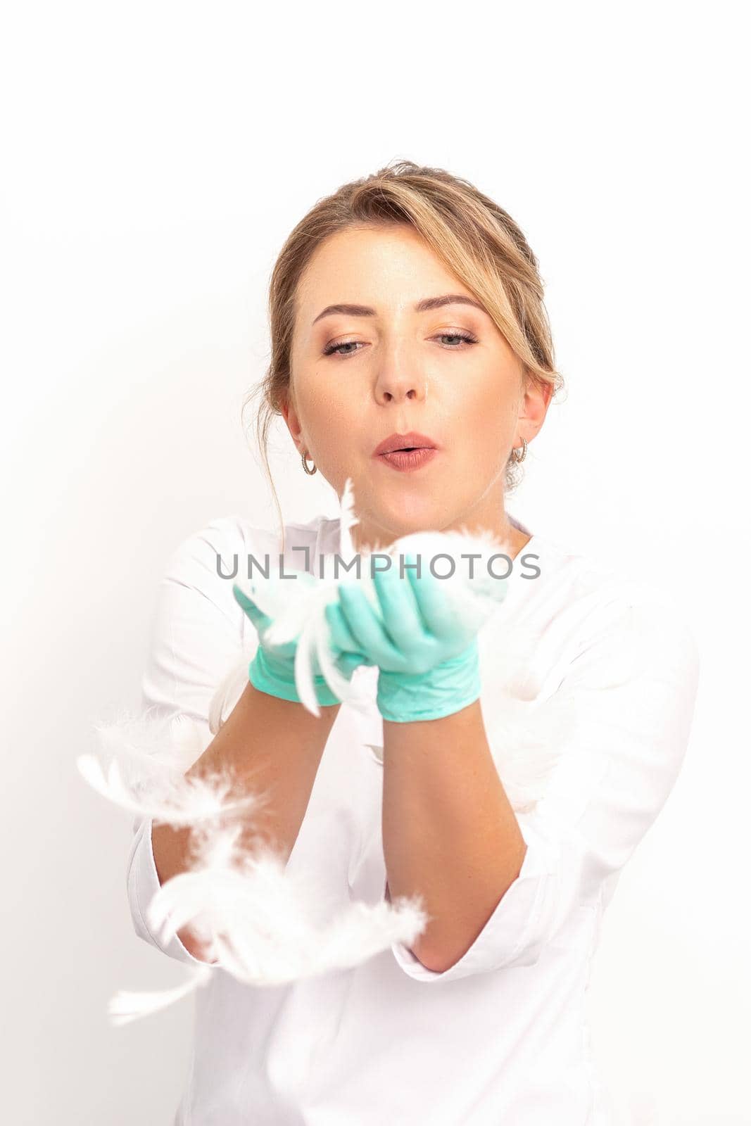 Young beautiful woman beautician in protective green gloves standing and blowing on feathers over white wall background. by okskukuruza