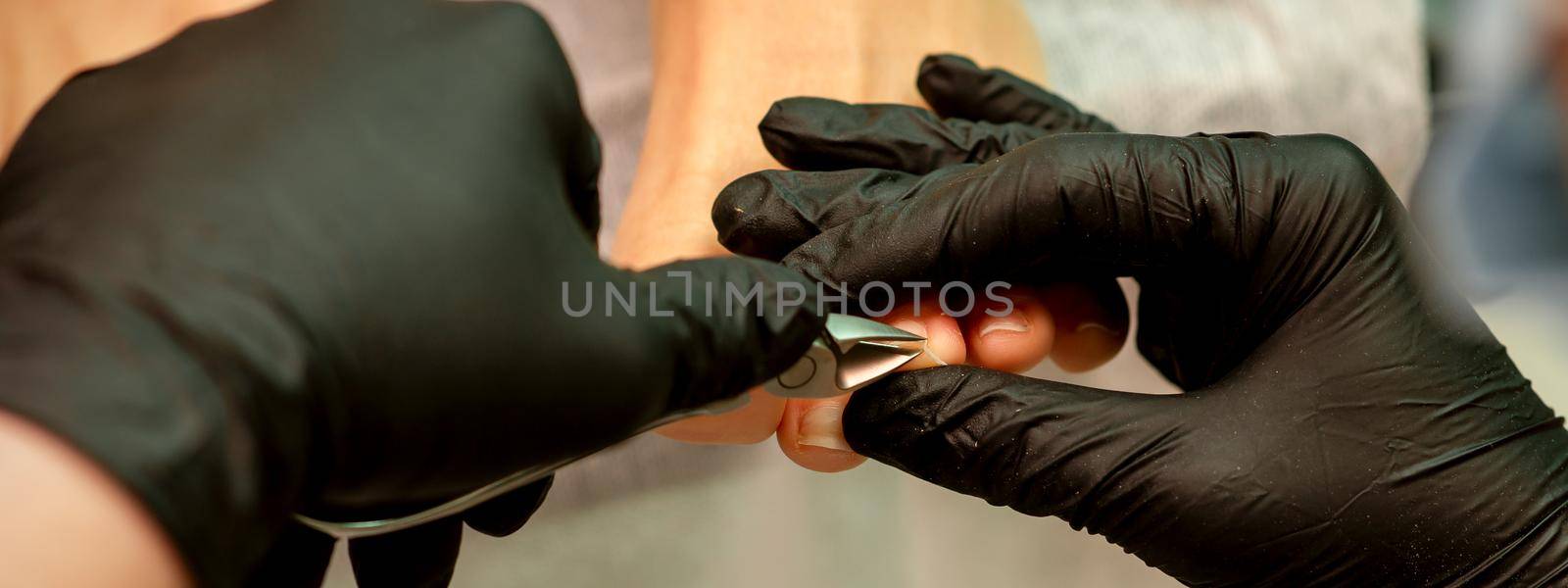Professional pedicure. Pedicure master wearing latex gloves cuts female toenails in the beauty salon, closeup. by okskukuruza