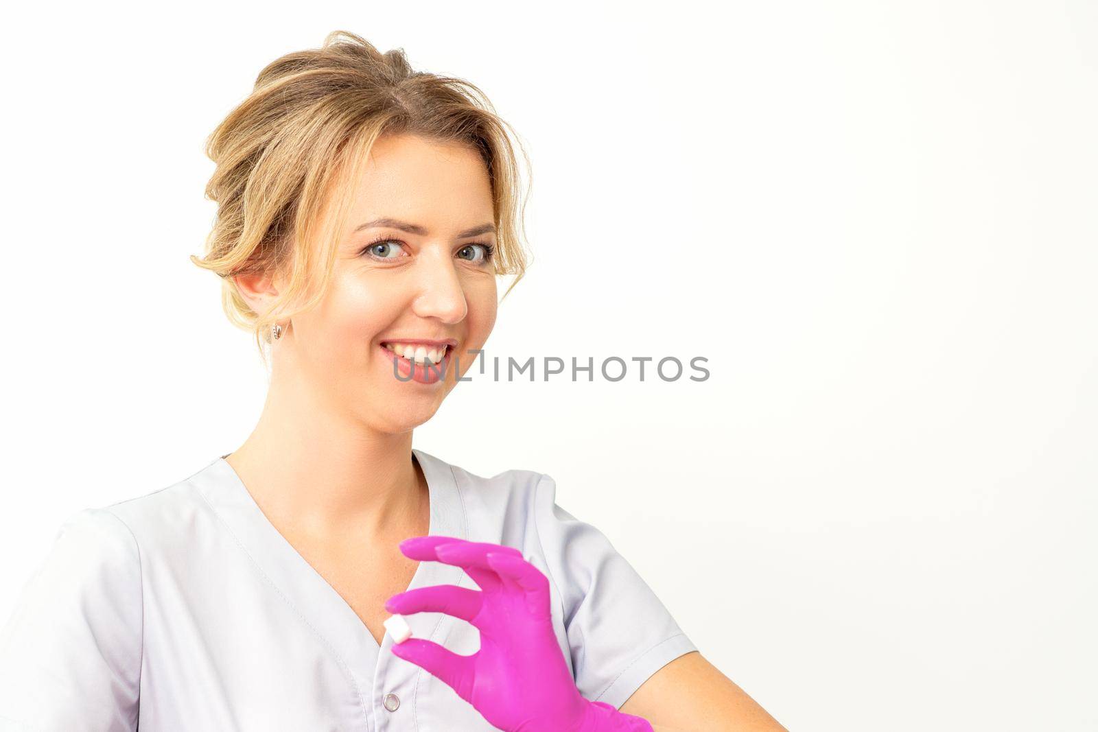 Portrait of a young smiling Caucasian beautician wearing pink gloves holding sugar cubes showing and looking at the camera against a white background