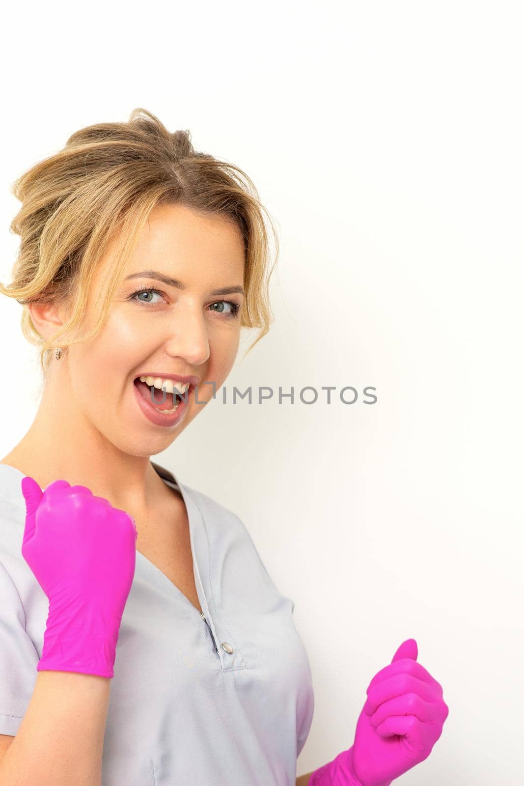 Happy caucasian woman doctor wearing pink gloves celebrates and raising fists on white background. by okskukuruza
