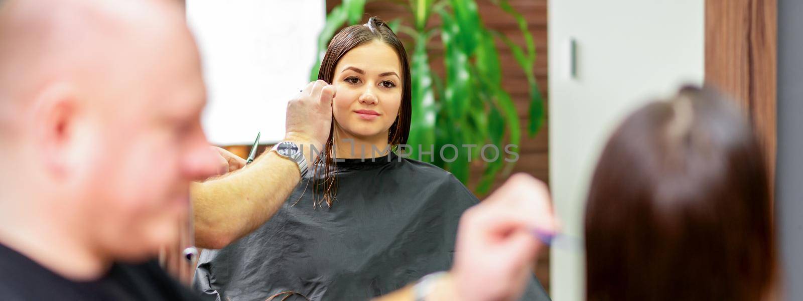 Reflection in the mirror of the young caucasian woman sitting and receiving haircut by male hairdresser at hairdresser salon. by okskukuruza