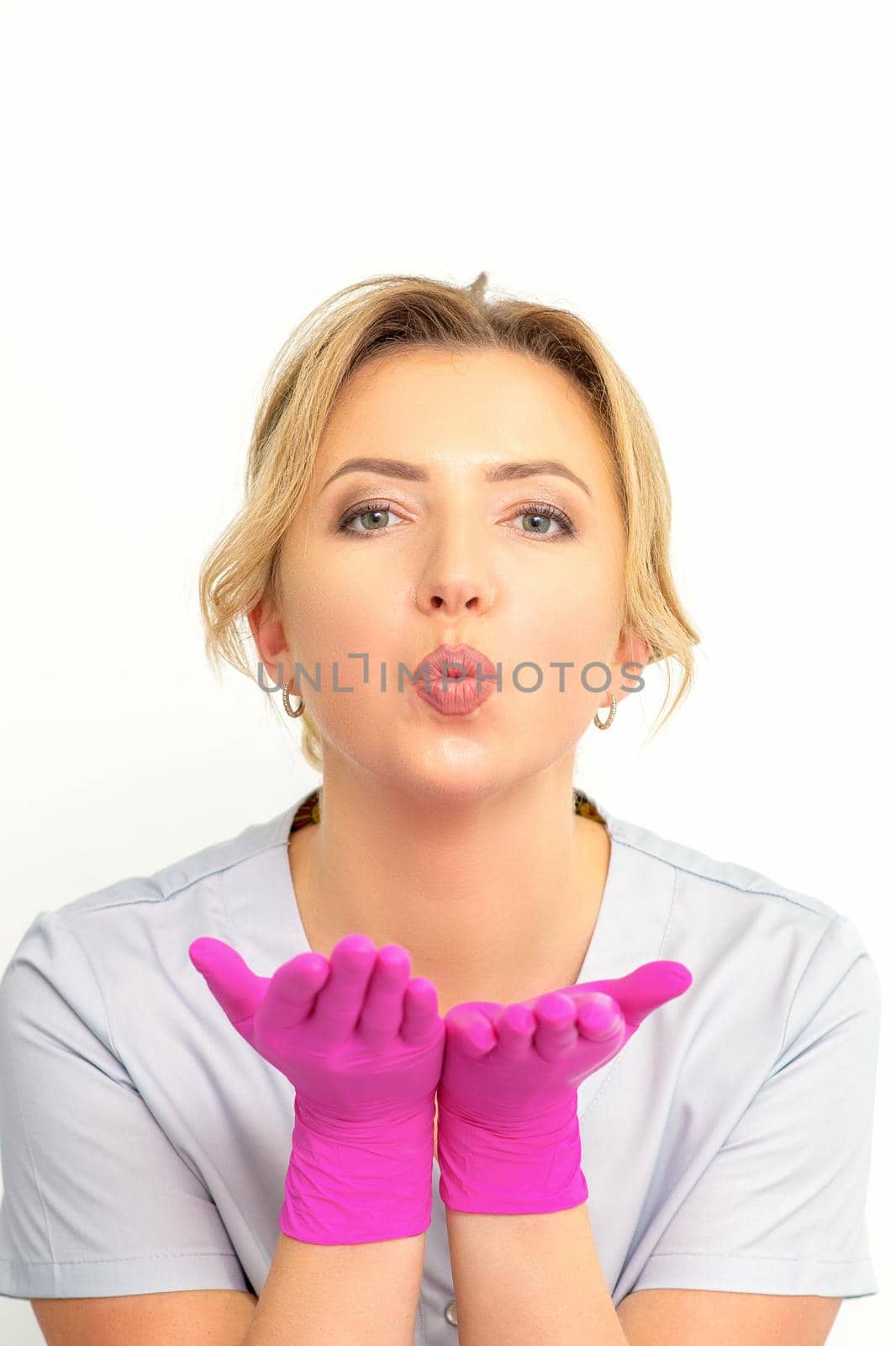 Young Caucasian doctor woman wearing pink gloves folding lips and holding palms to send air kiss isolated white background. by okskukuruza