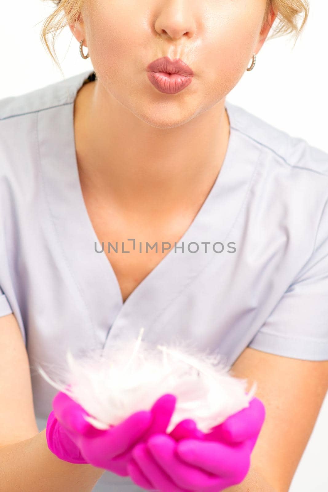 Young beautiful woman beautician in protective pink gloves standing and holding white feathers over white wall background. by okskukuruza