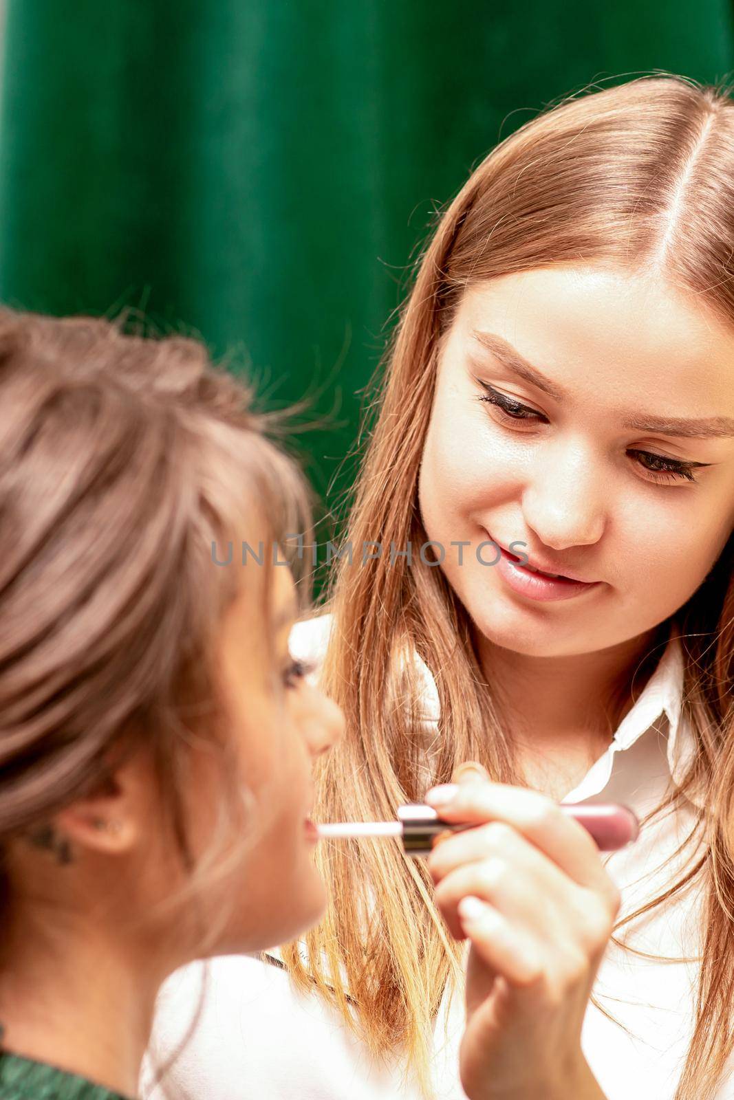 Makeup in the process. The makeup artist applies pink gloss lipstick on the lips of the beautiful face of the young caucasian woman in a beauty salon