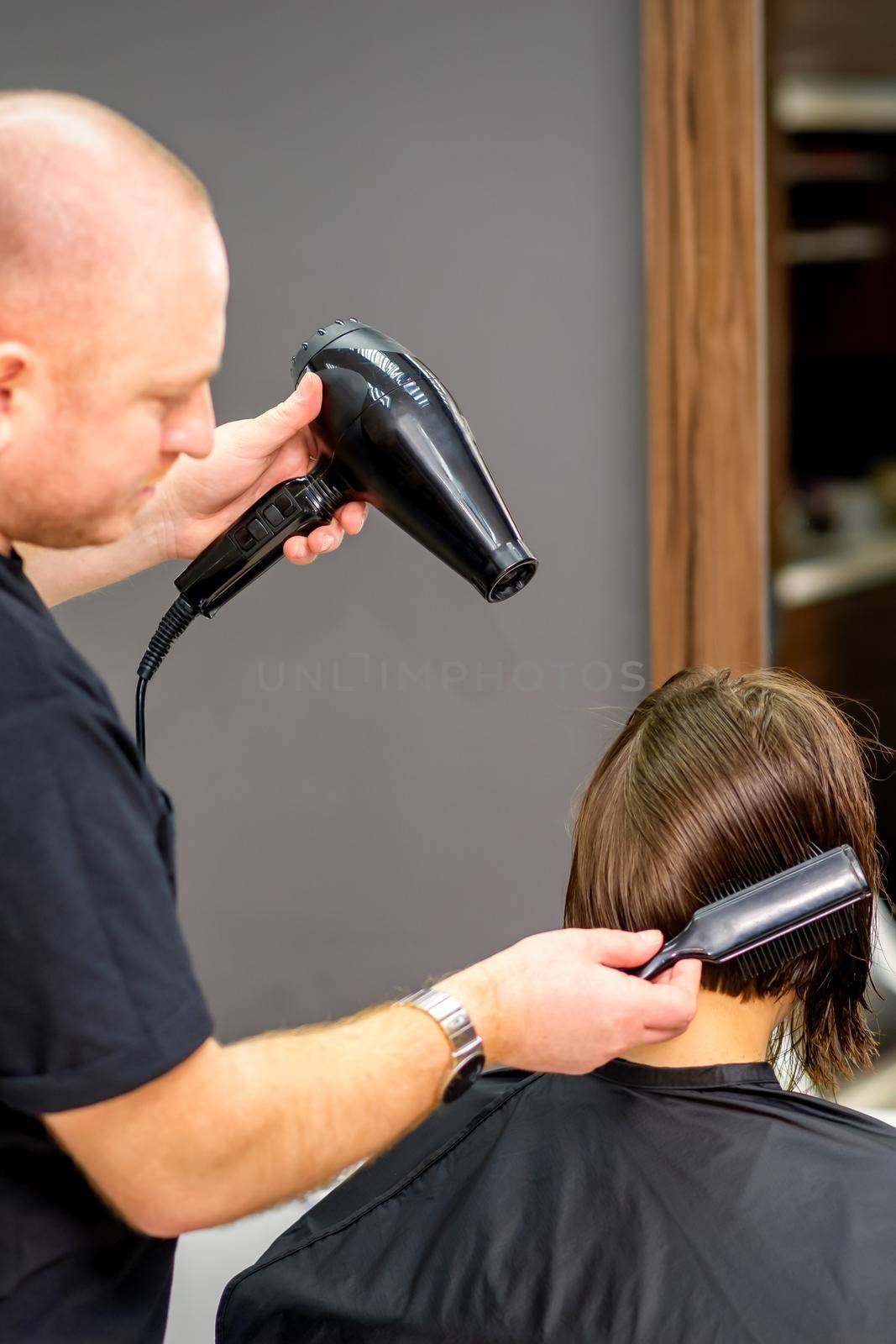 Male hairdresser drying short hair of young caucasian brunette woman with a black hairdryer and black round brush in a hairdresser salon