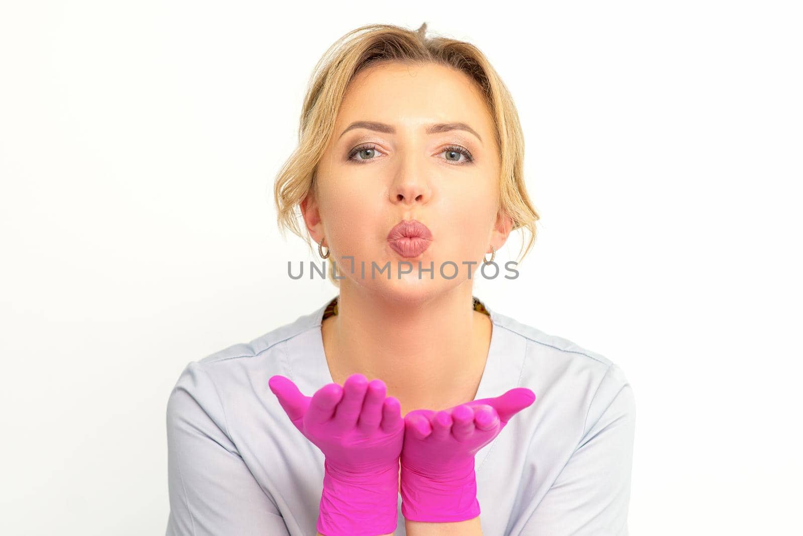 Young Caucasian doctor woman wearing pink gloves folding lips and holding palms to send air kiss isolated white background