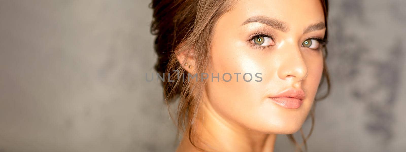 The fashionable young woman. Portrait of the beautiful female model with long hair and makeup. Beauty young caucasian woman with a brown curly hairstyle on the background of a gray wall