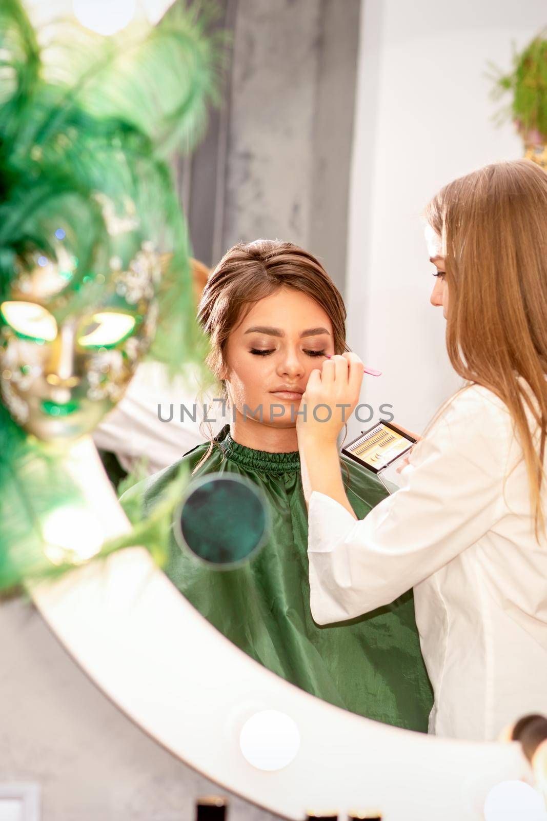 Professional makeup artist applies makeup to the eyes of the beautiful young woman in the mirror smiling in a beauty salon