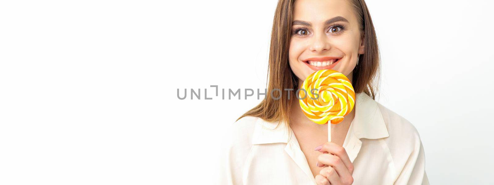Beautiful young caucasian woman wearing a white shirt licking a lollipop on a white background. by okskukuruza